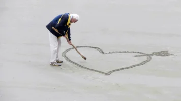 Senior man drawing heart with an arrow with a wooden stick in sand on the beach.