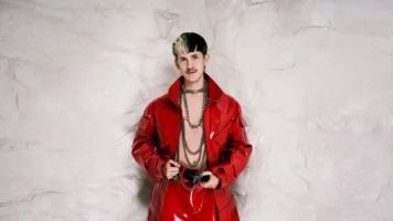 Picture of Teeo with a white wall behind him. He is in red lacquer - and his jacket is open and are showing skin.