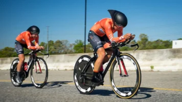 To female are cycle racing - both of them has cycle helmet, black cycle shorts and orange shirts