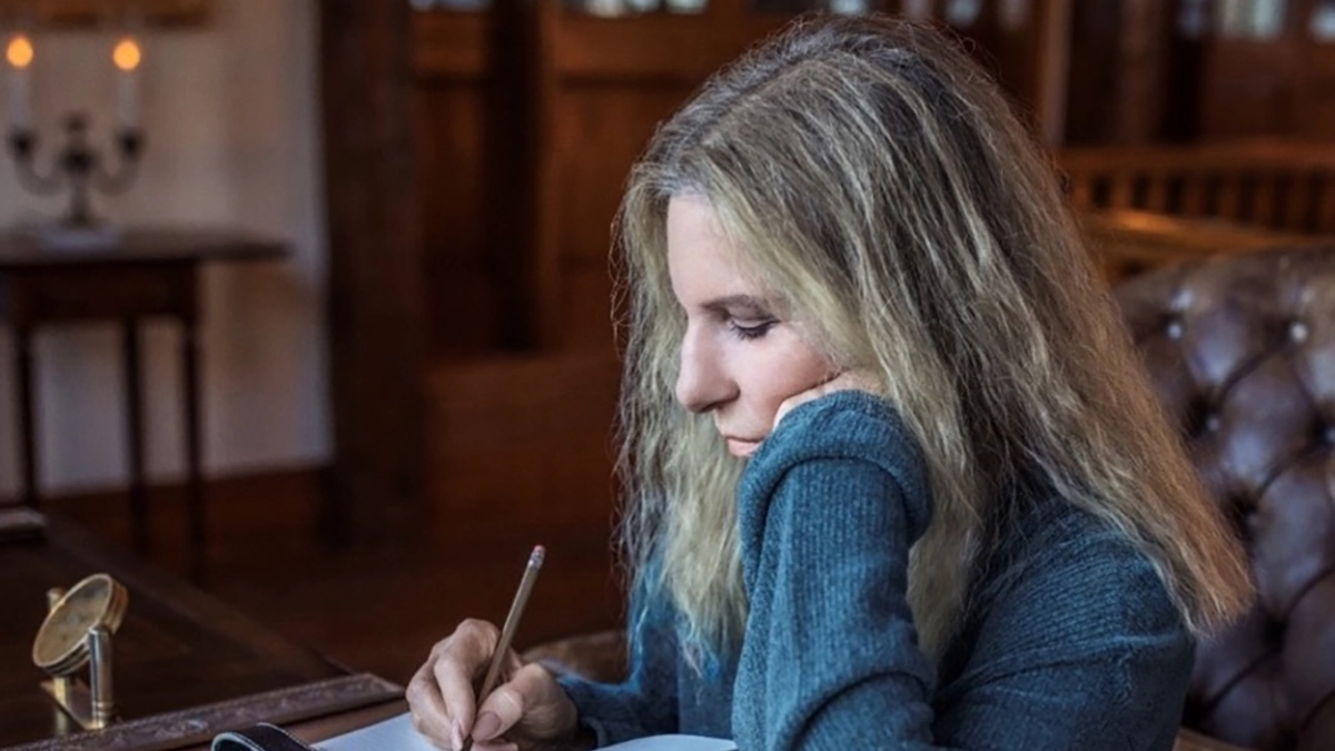 Barbra Streisand sits at a desk, using a pen to write. She gazes down at her notebook, wearing a grey sweater.